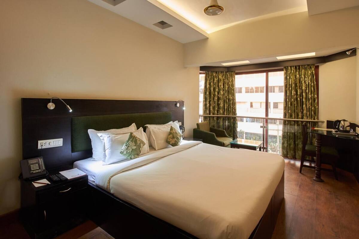 A hotel room with a large bed, bedside table with a phone, a desk with a chair, and a view of buildings through the window.