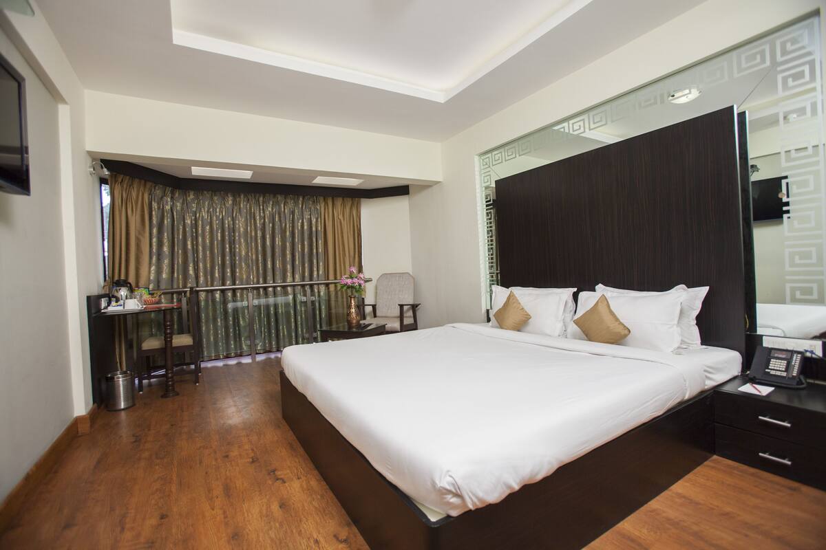 A hotel room with a large bed, a desk with a chair, a small table with a vase, and a mirror on the wall.