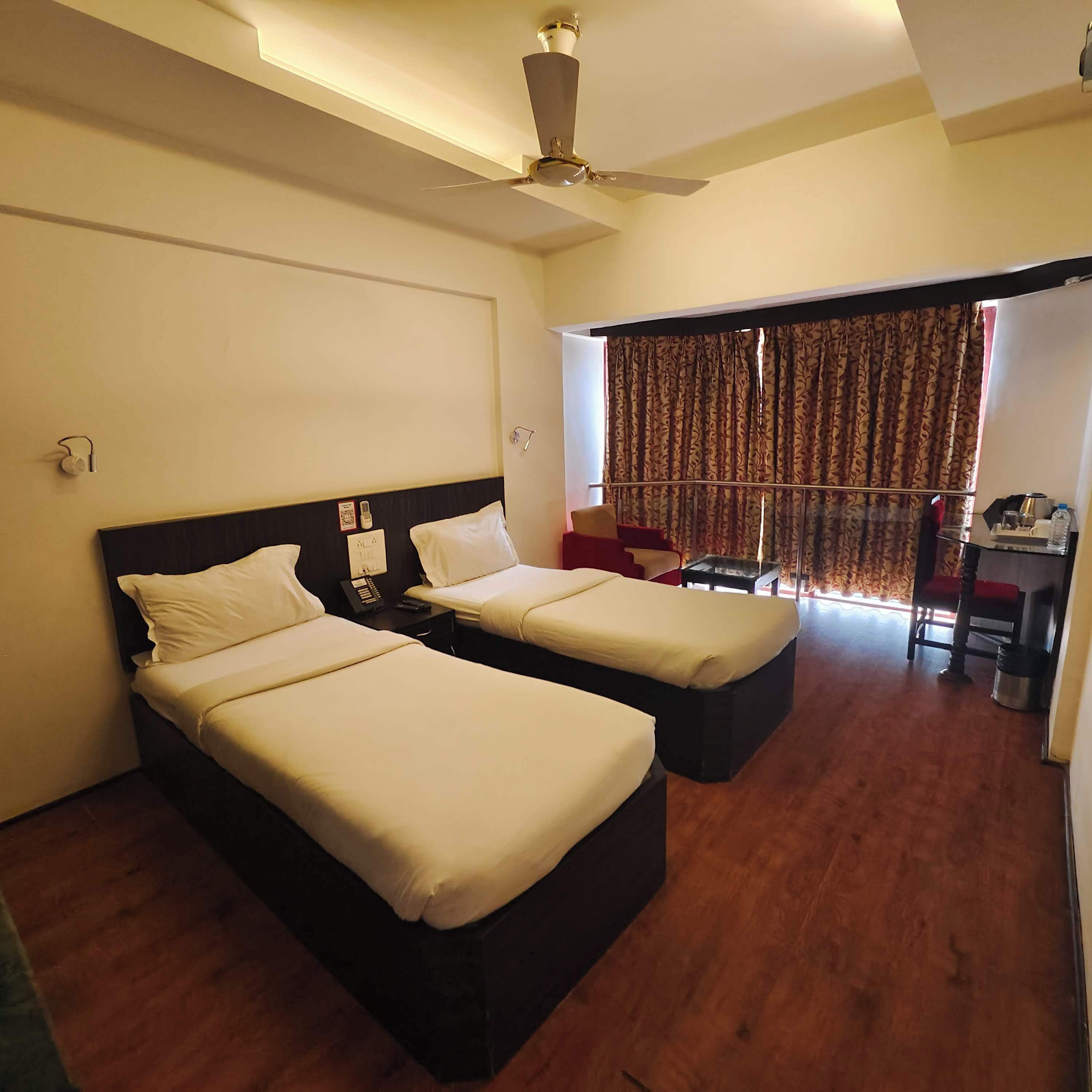 A hotel room with two beds, a ceiling fan, a desk with a chair, and a window with curtains.