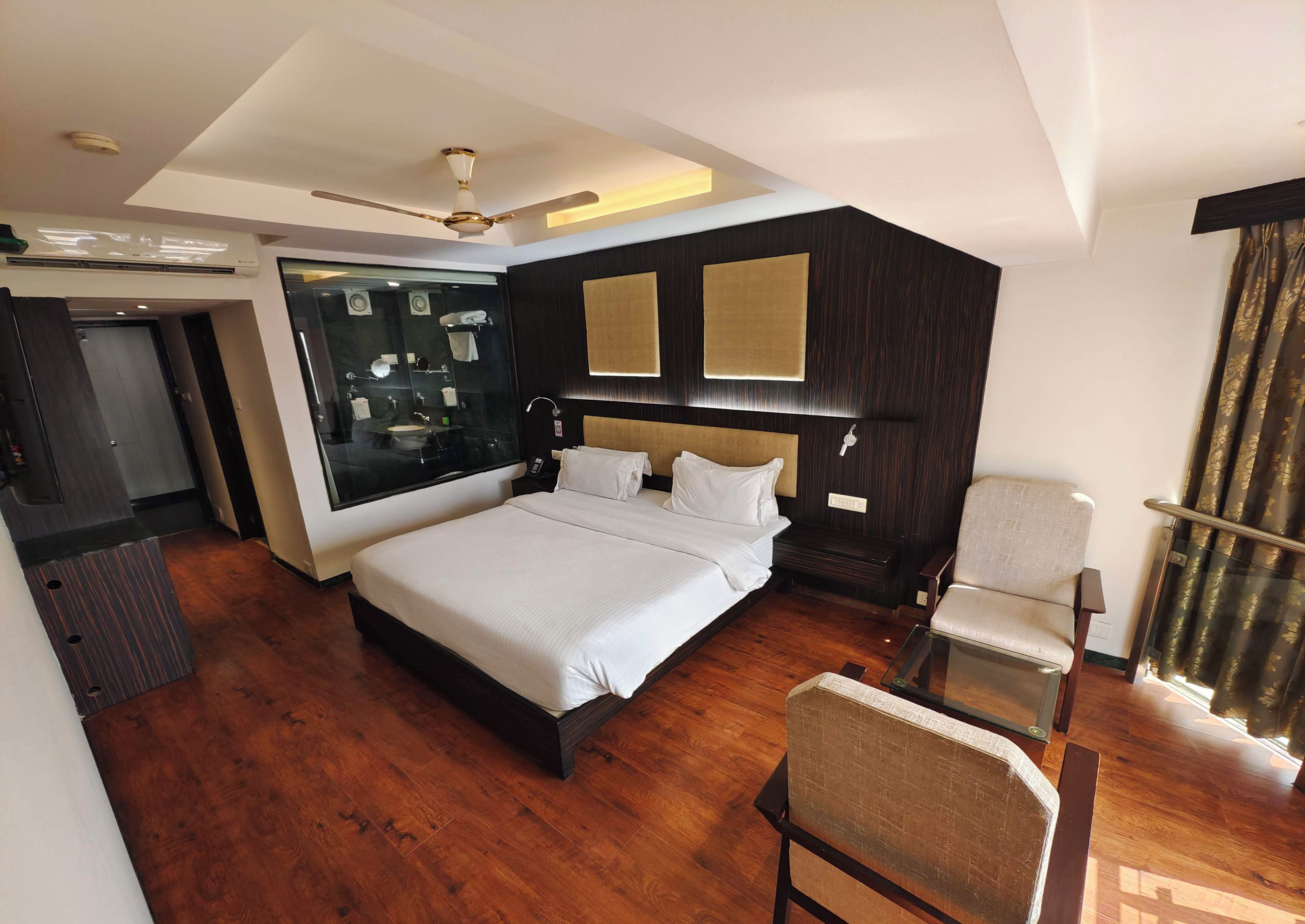 A modern hotel room with a large bed, a glass-enclosed bathroom, a desk, and a chair.
