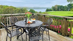 Een ronde metalen tafel met stoelen op een houten terras, met uitzicht op een meer en een groen landschap.