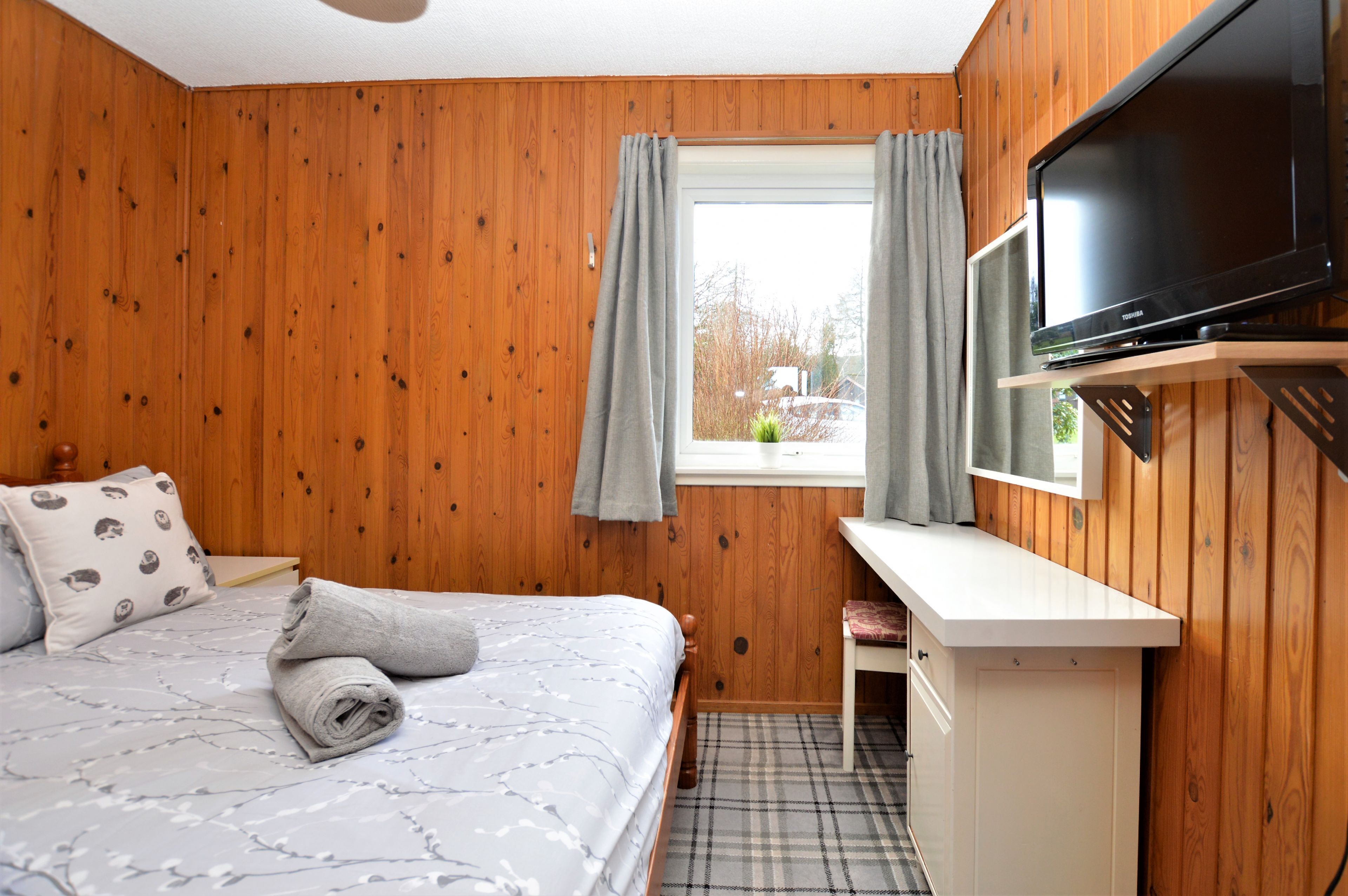 A wooden-paneled room with a bed, a TV mounted on the wall, and a window with curtains.