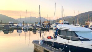 A marina with boats docked at wooden piers, calm water, and a mountainous backdrop.