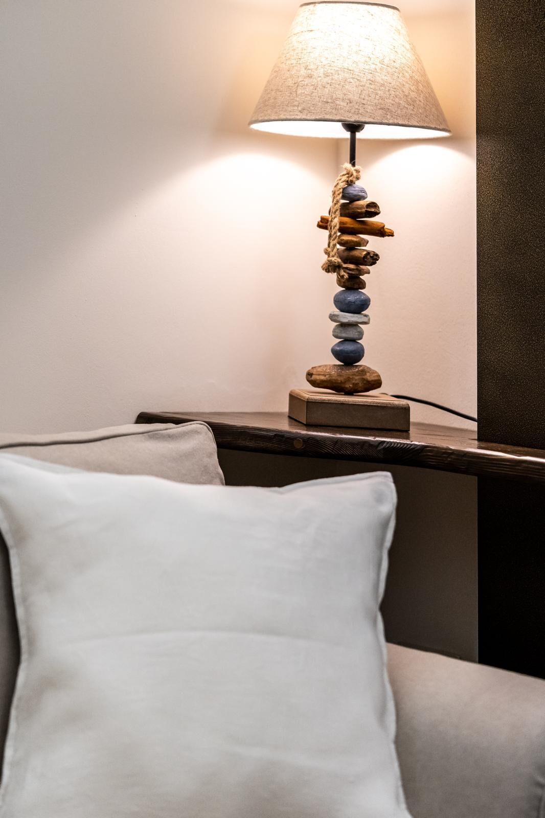 A bedside table with a lamp featuring a stack of stones and a rope, a white pillow, and a wooden nightstand.