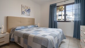 Een slaapkamer met een groot bed, een nachtkastje, een dressoir, een raam met gordijnen, en een wandlamp.