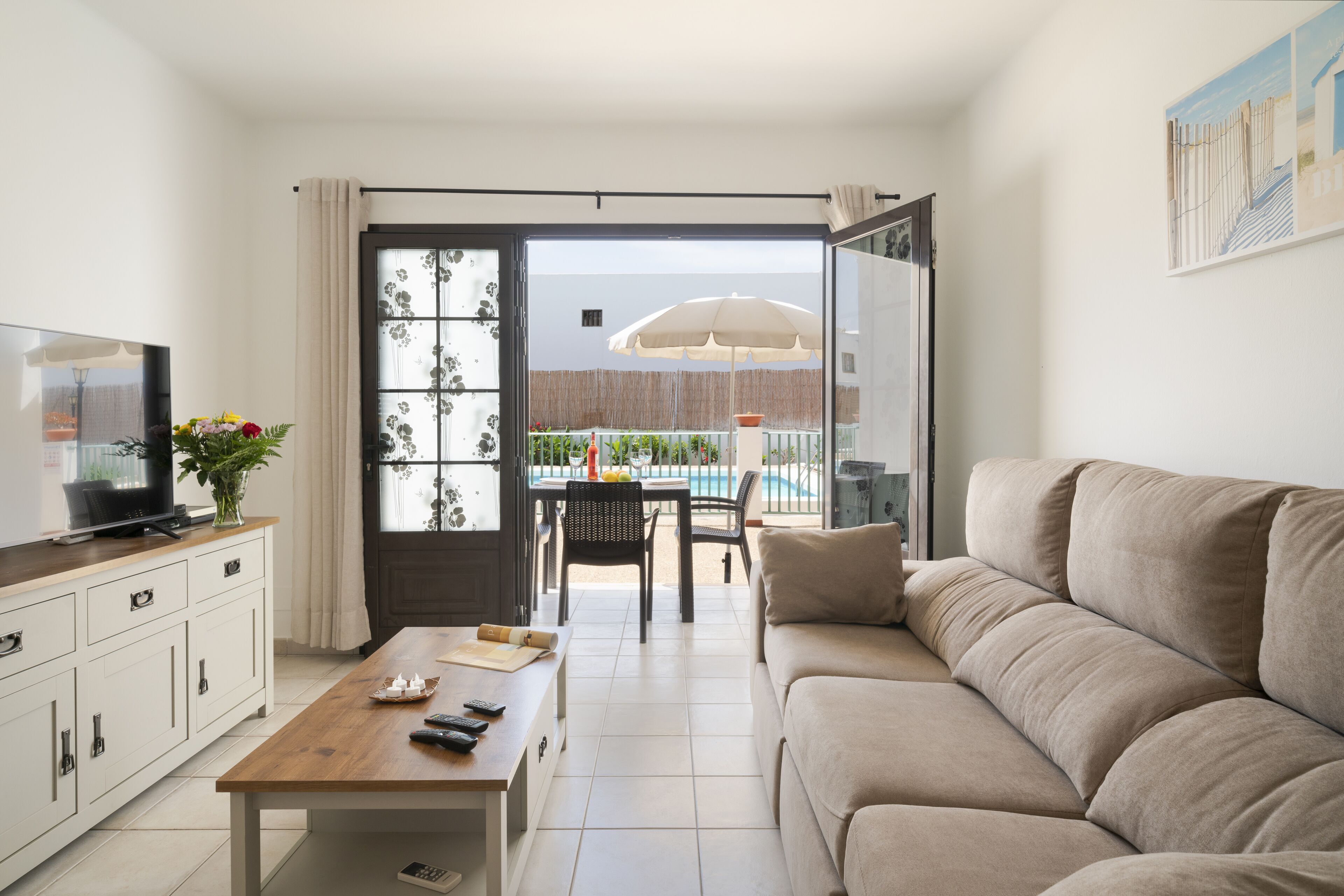 Een woonkamer met een bank, salontafel, televisie en een eethoek met stoelen en een parasol.