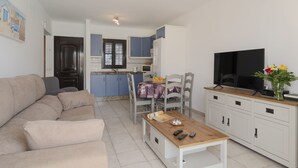 Een woonkamer met een bank, een salontafel en een televisie. Er is een keukenhoek met kastjes en een raam.