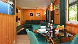 Uma área de refeições com mesa de vidro, cadeiras verdes e uma parede de madeira com quadros emoldurados.