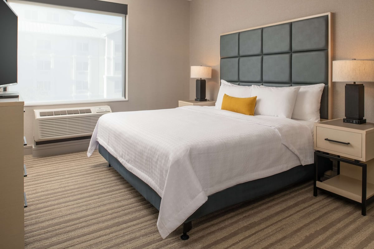 A hotel room with a large bed, two bedside tables with lamps, a window with blinds, and a radiator.