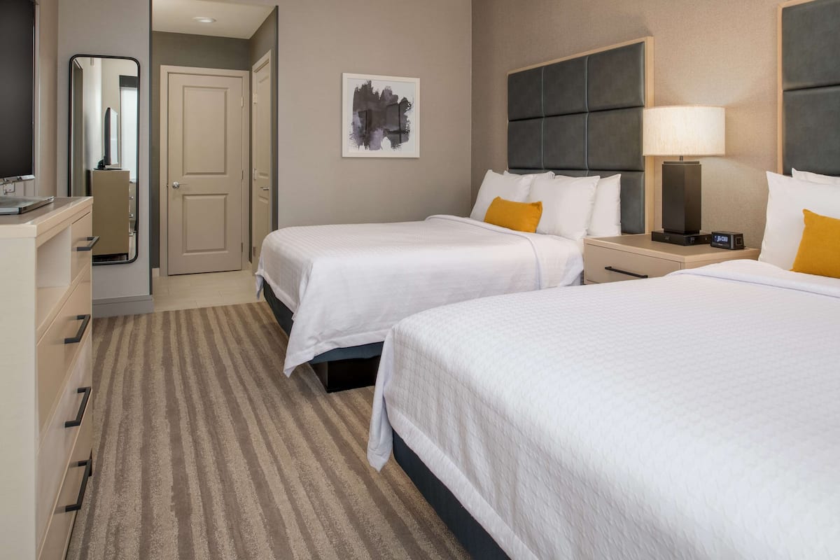 A hotel room with two beds, a bedside table with a lamp, a nightstand, a large mirror, and a framed artwork on the wall.
