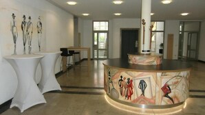 A spacious lobby with a central round table, white tables, and chairs, and a mural on the wall. - Kolping Hotel Schweinfurt (Schweinfurt)