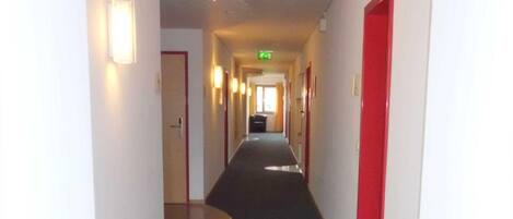 Un largo pasillo estrecho con alfombra oscura, paredes blancas y puertas rojas a ambos lados.