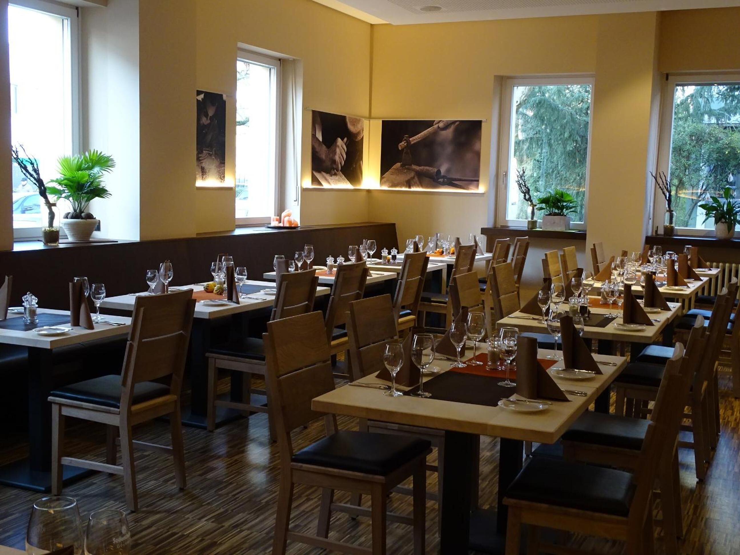Ein Restaurant mit mehreren Esstischen, Holzstühlen und Blumentöpfen an den Fensterbänken.