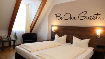 Quarto de hotel com cama de casal, cabeceira de madeira, parede com a inscrição "Be Our Guest...", duas luminárias de cabeceira, uma mesinha com uma planta e uma janela com cortinas.