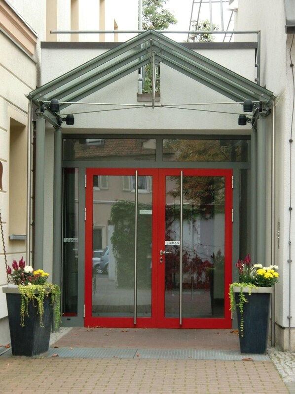 A building entrance with a red door and a glass panel, flanked by two flower planters. - Kolping Hotel Schweinfurt (Schweinfurt)