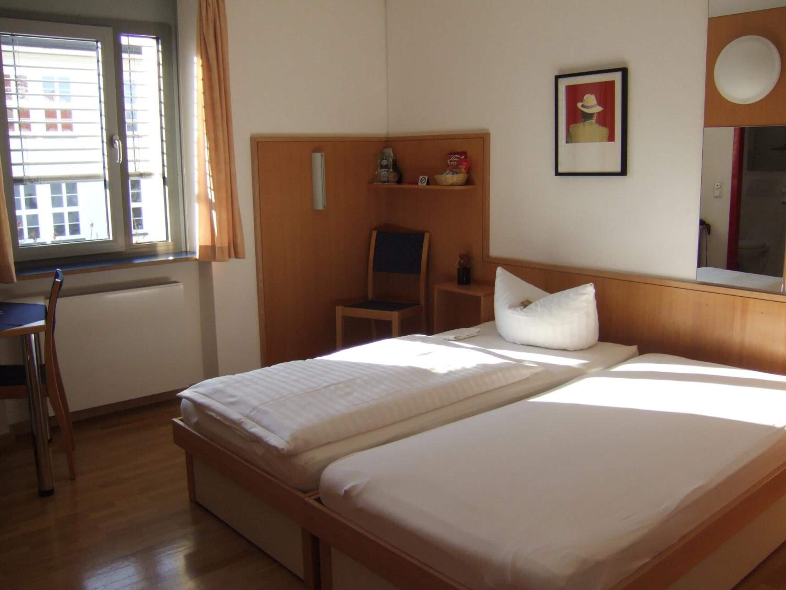 Habitación de hotel con dos camas, un escritorio, una silla, un estante con artículos de aseo y una ventana con cortinas.