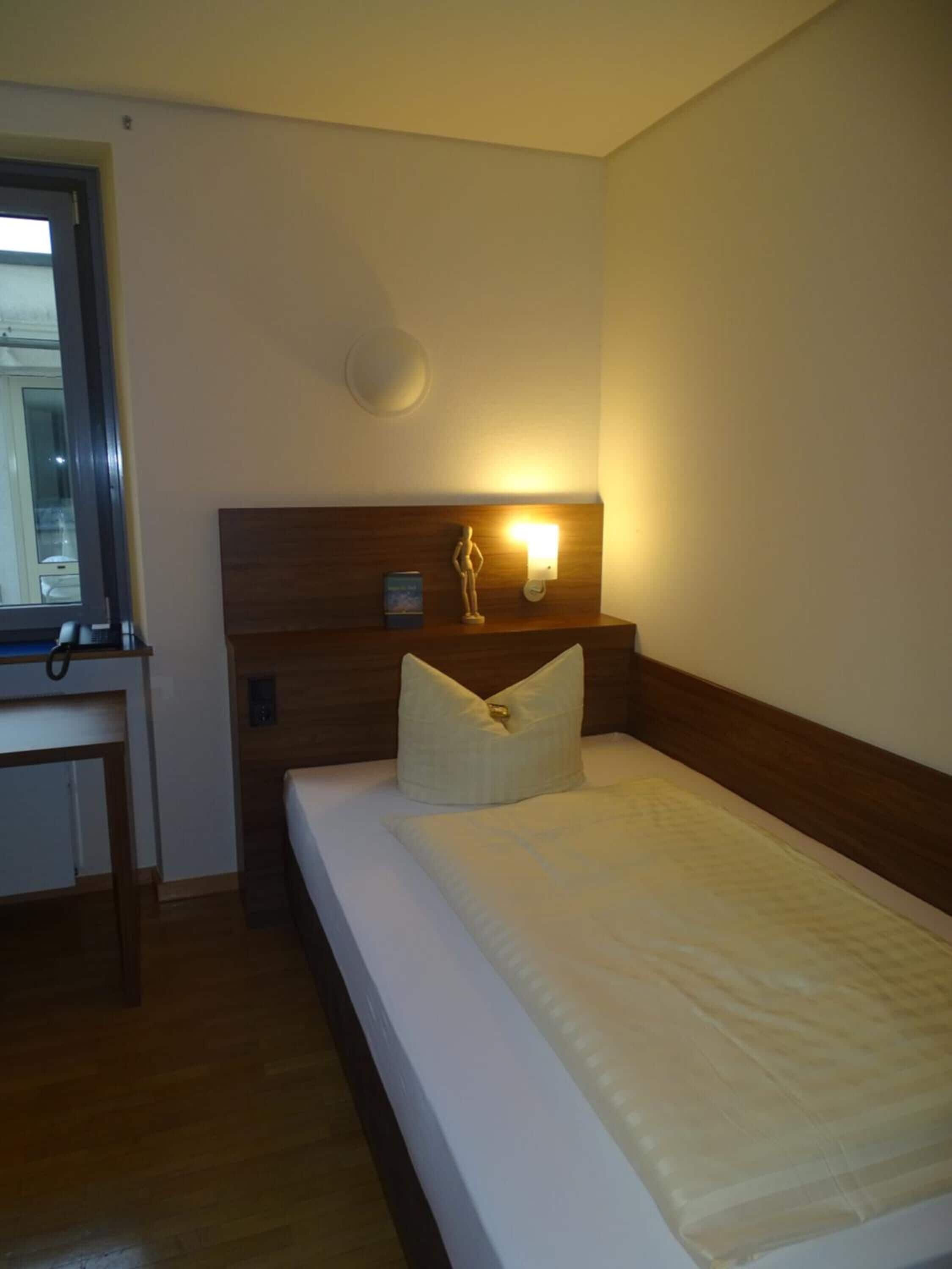 A hotel room with a wooden headboard, a bedside table, a nightstand, and a wall-mounted lamp.