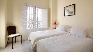 A room with two beds, a chair, a window with shutters, a lamp, and a framed picture on the wall.