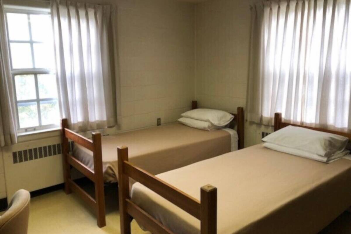 A small, simple room with two single beds, a chair, and a window with curtains.