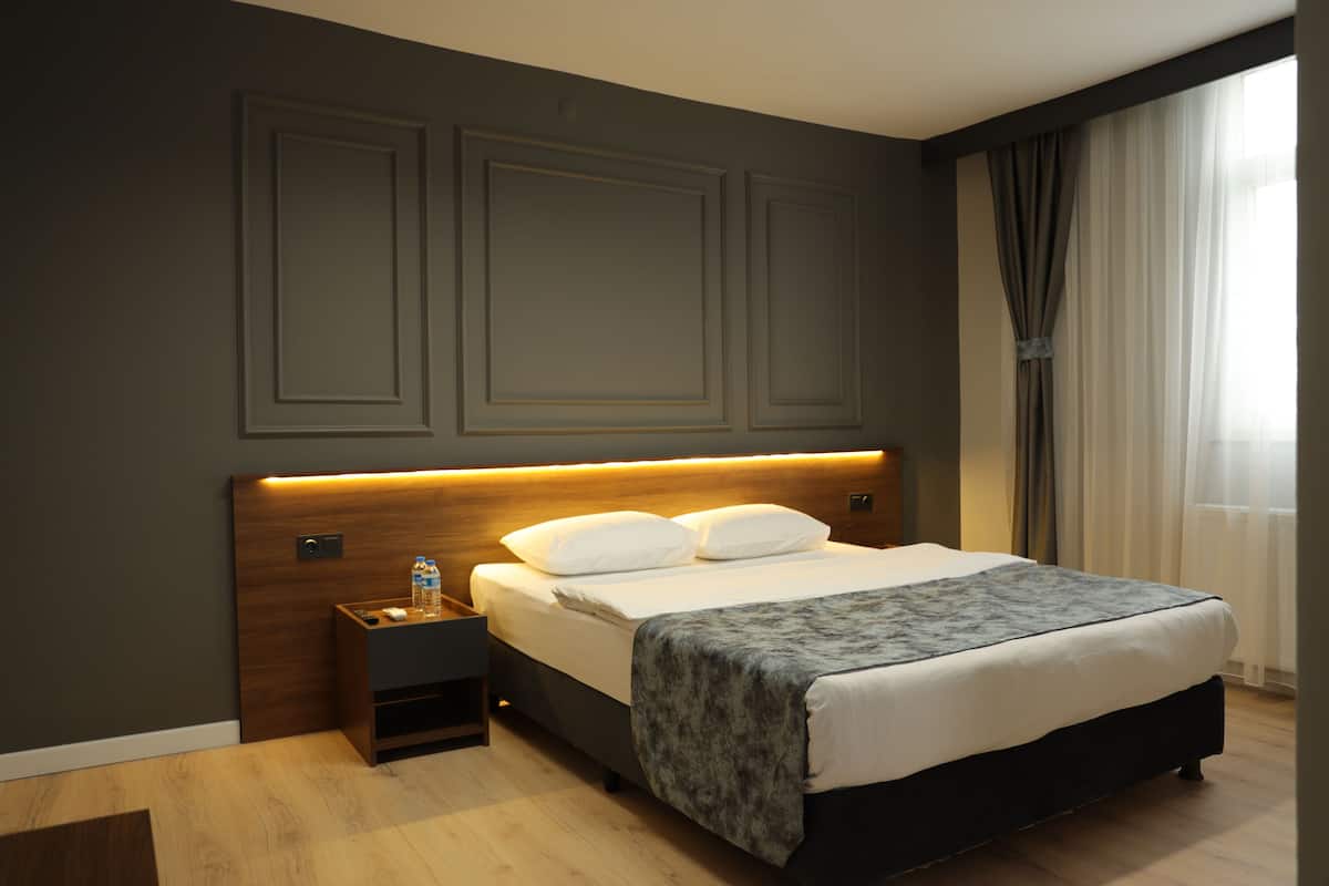 A modern hotel room with a wooden headboard, a neatly made bed, a nightstand with a bottle, and a window with curtains.