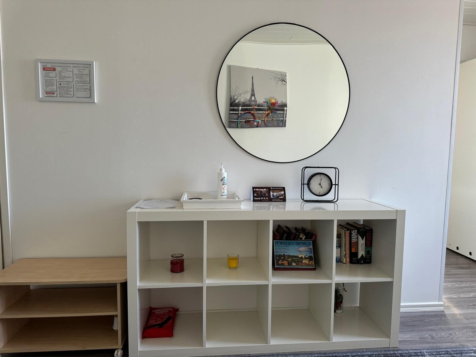 A white console table with a round mirror, a clock, and a small shelf with books and a candle.