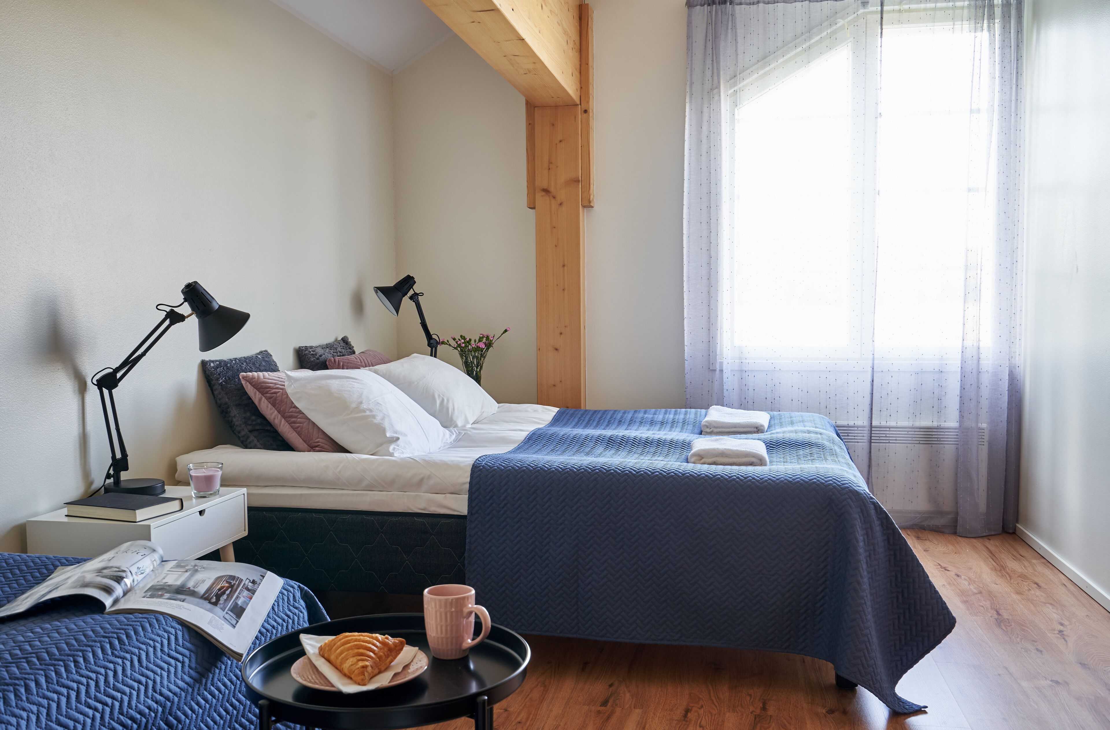 Un dormitorio con dos camas, una mesita de noche con lámparas, una pequeña mesa con una bandeja de comida y una ventana con cortinas.