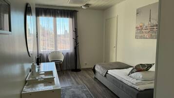 Ein Schlafzimmer mit Bett, Schreibtisch, Stuhl, einem Fenster mit Vorhängen, einem Kleiderständer und einem Bild des Eiffelturms.