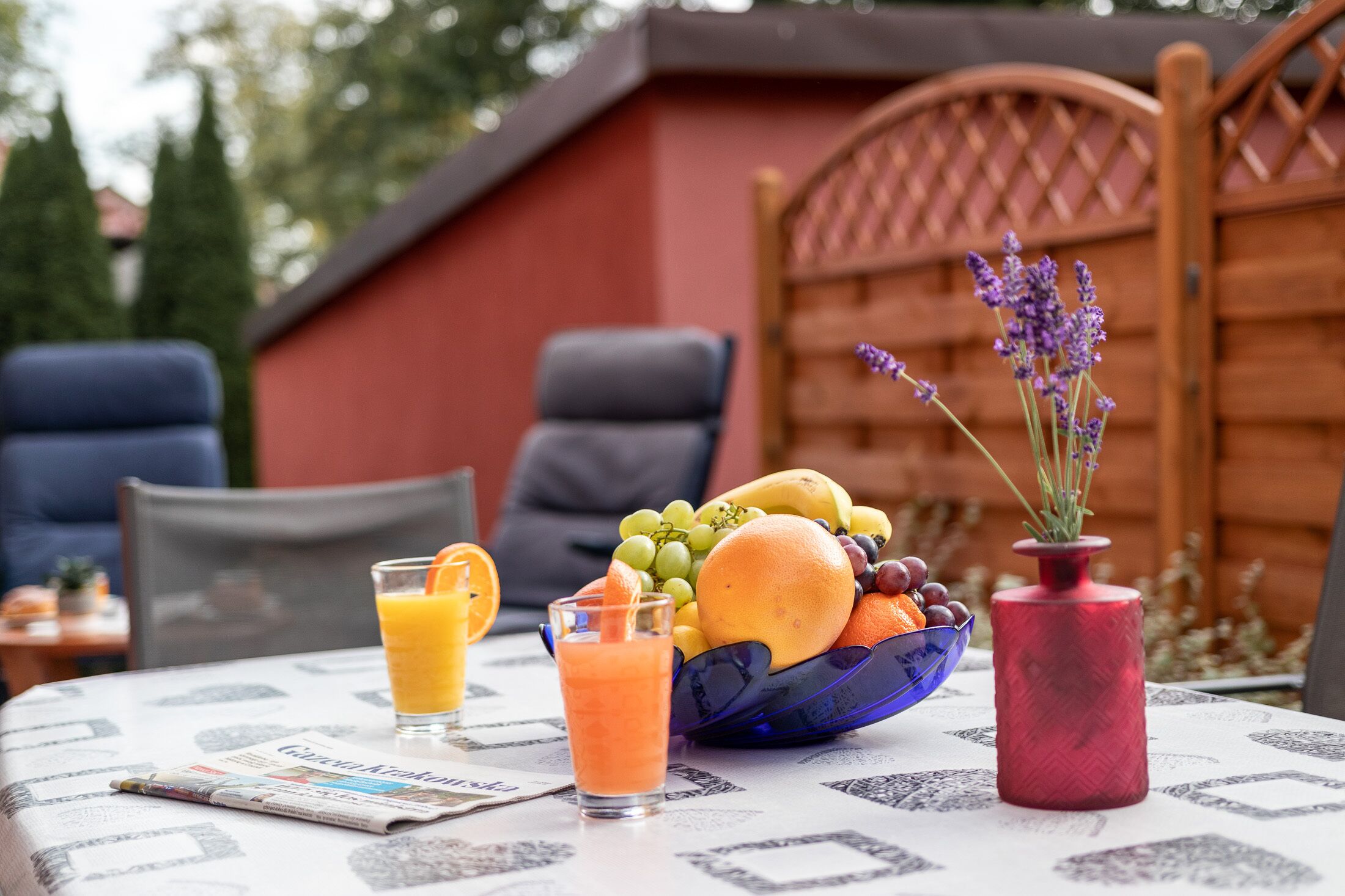 Una mesa con un frutero, un vaso de jugo de naranja, un florero rojo con lavanda y un periódico.