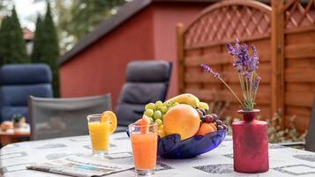 Una mesa con un frutero, un vaso de jugo de naranja, un florero rojo con lavanda y un periódico.