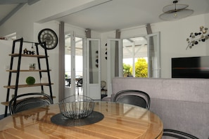 A modern dining area with a round wooden table, metal chairs, a black metal bowl, a ladder shelf with decorative items, a wall clock, and a television on the right. - Magnificent villa close to the sea (Le Diamant)