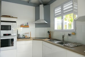 Una cocina moderna con armarios blancos, un microondas Bosch, horno empotrado, pileta de acero inoxidable y una ventana con contraventanas blancas.