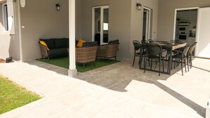 Ein überdachter Patio-Bereich mit Esstisch und Stühlen, einer Couch mit Kissen sowie einem durch eine offene Tür einsehbaren Küchenbereich.