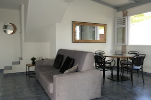 Una sala de estar moderna con un sofá gris, un espejo redondo, una mesa de comedor con sillas y una ventana con persianas.