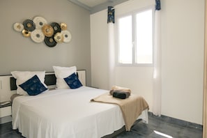 A bedroom with a bed, pillows, a bedside table, a wall art piece, a window, and a curtain. - Magnificent villa close to the sea (Le Diamant)