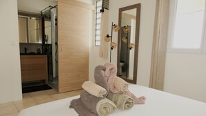 A neatly made bed with rolled towels, a wooden door, a bathroom with a mirror, and a window. - Magnificent villa close to the sea (Le Diamant)