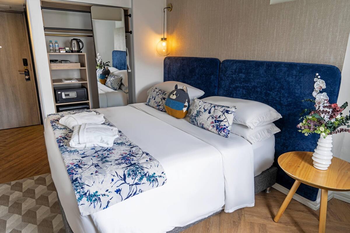 A neatly made bed with a floral bedspread, a blue headboard, and a bedside table with a vase of flowers.
