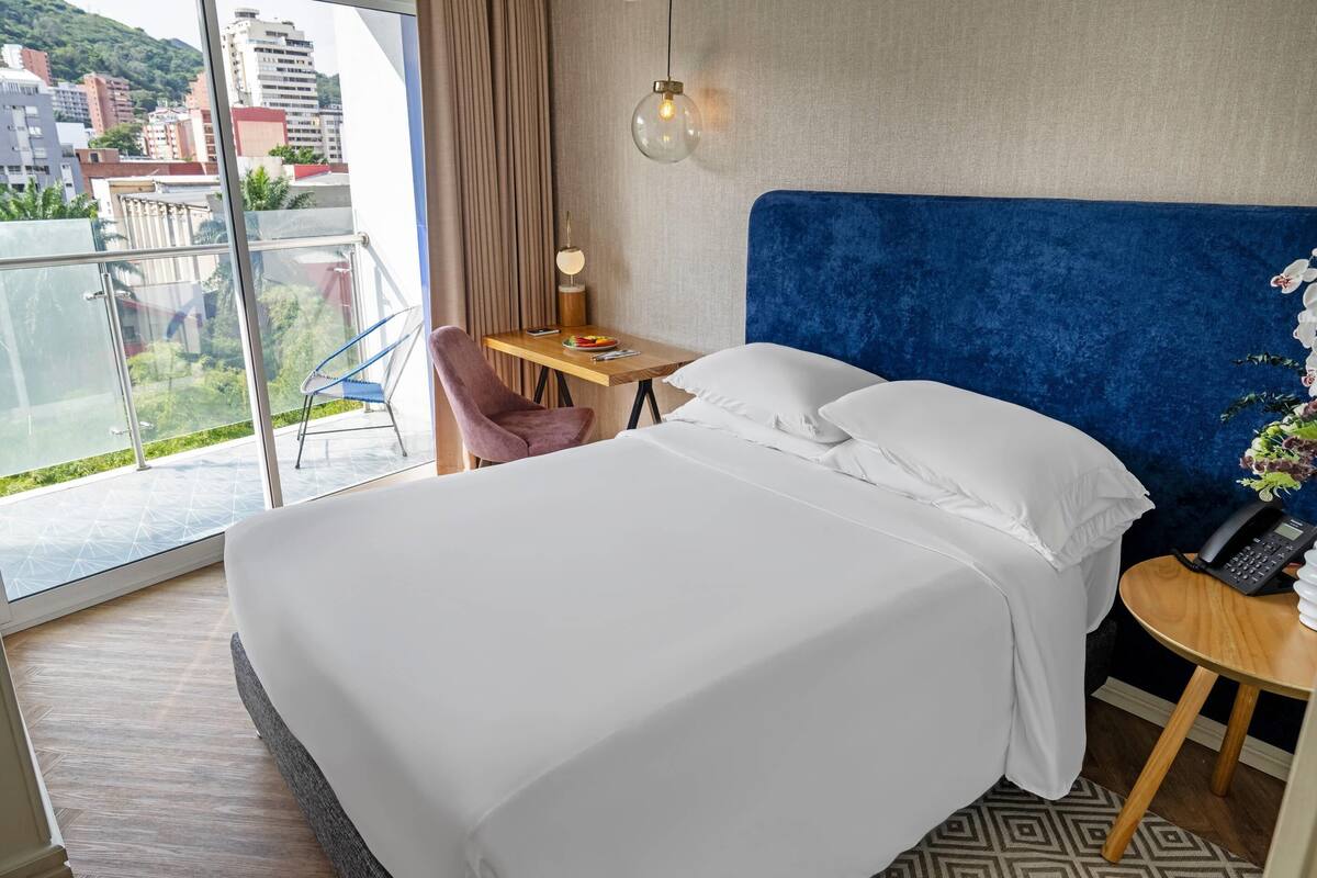 A hotel room with a large bed, a desk with a lamp, a chair, and a view of the city through large windows.