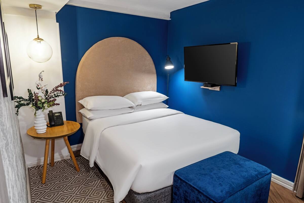 A bedroom with a bed, a round headboard, a bedside table with a lamp, a TV, and a blue ottoman.