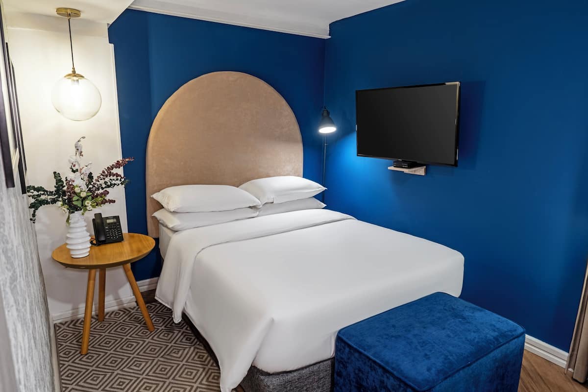A bedroom with a bed, a round headboard, a bedside table with a lamp, a TV, and a blue ottoman.