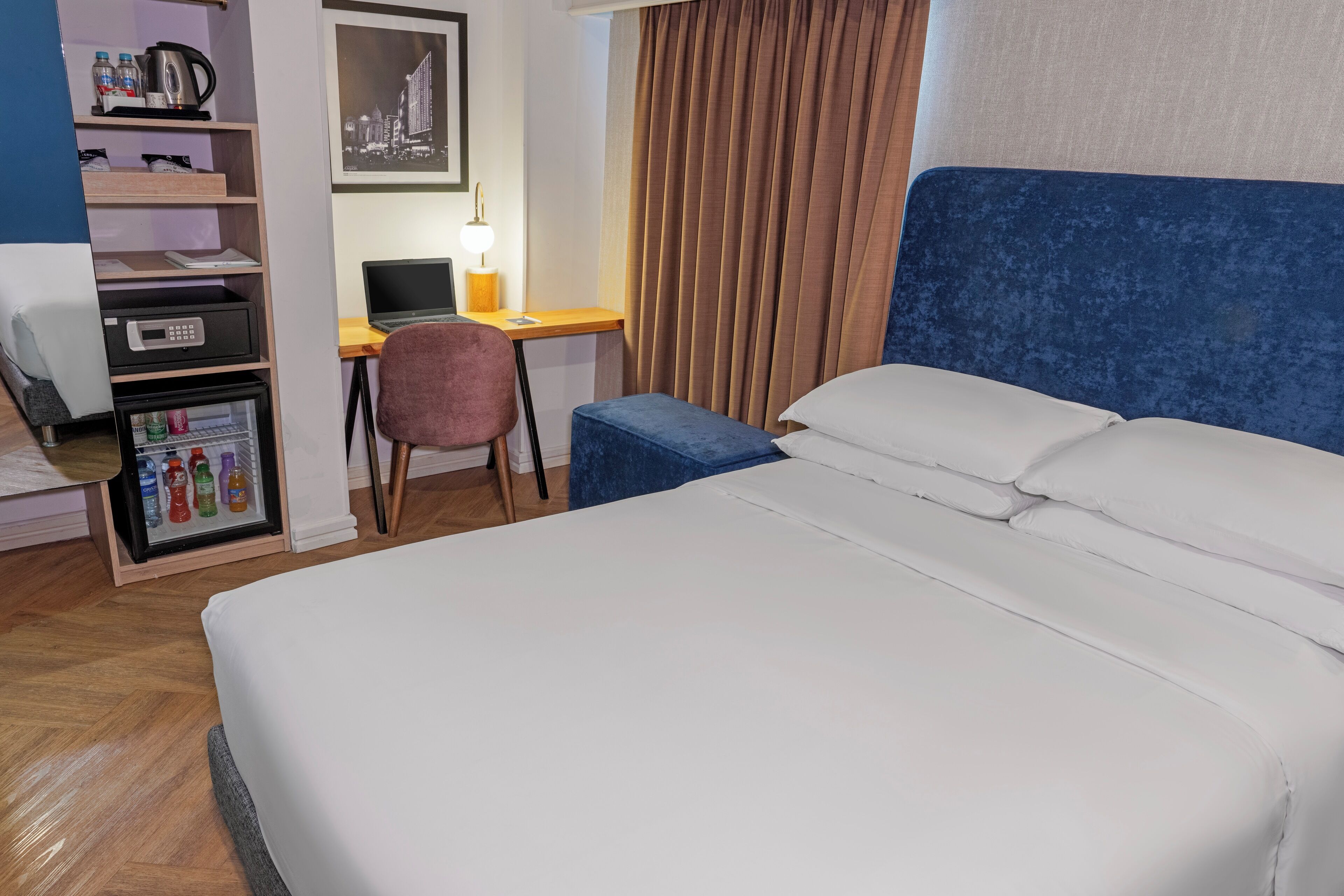 A hotel room with a bed, a desk with a laptop, a chair, a mini-fridge with beverages, and a coffee maker.