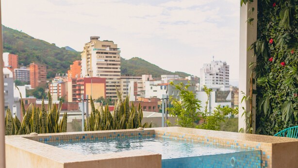 Una piscina en la azotea con borde de azulejos, un cartel que indica un jacuzzi y vistas de un paisaje urbano con edificios y colinas al fondo.