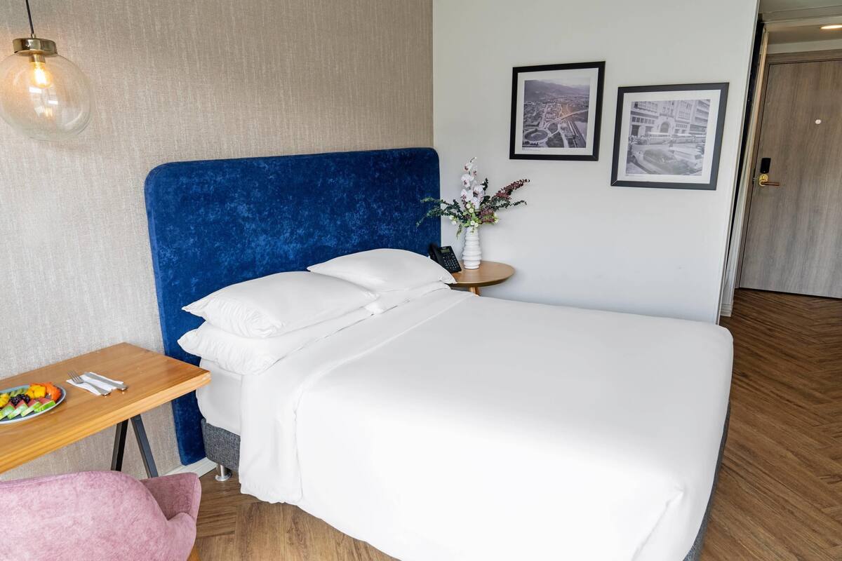 A hotel room with a blue headboard, white bedding, a wooden side table with a fruit plate, a pink chair, and two framed pictures on the wall.