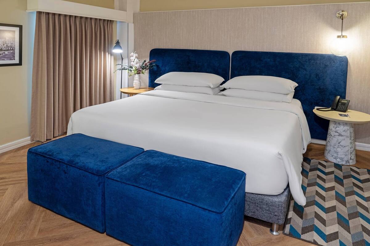 A hotel room with a large bed, a blue headboard, a bedside table with a lamp, a small round table with a phone, and a blue ottoman.