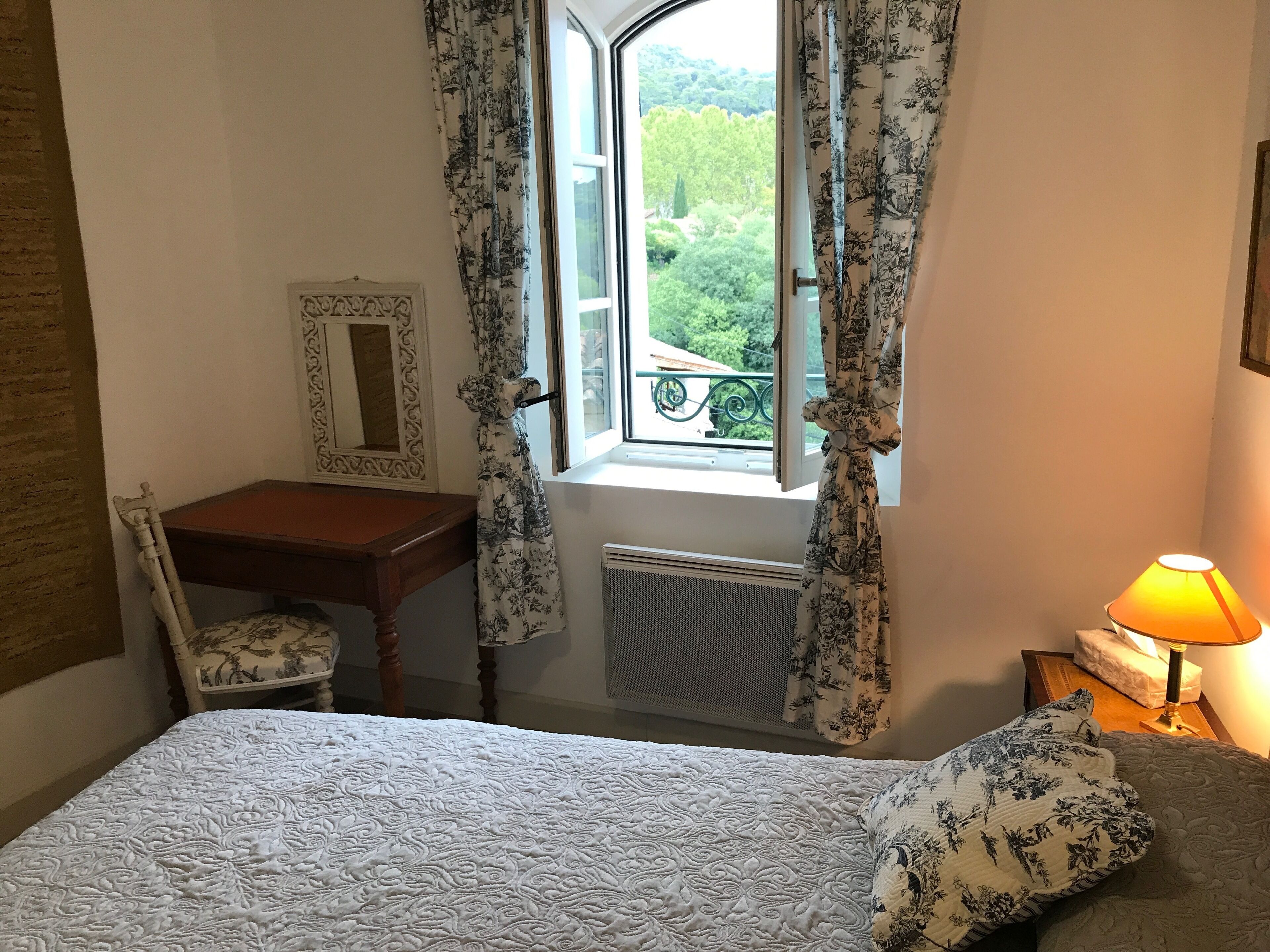A bedroom with a bed, a desk, a chair, a window with curtains, a radiator, and a bedside lamp.