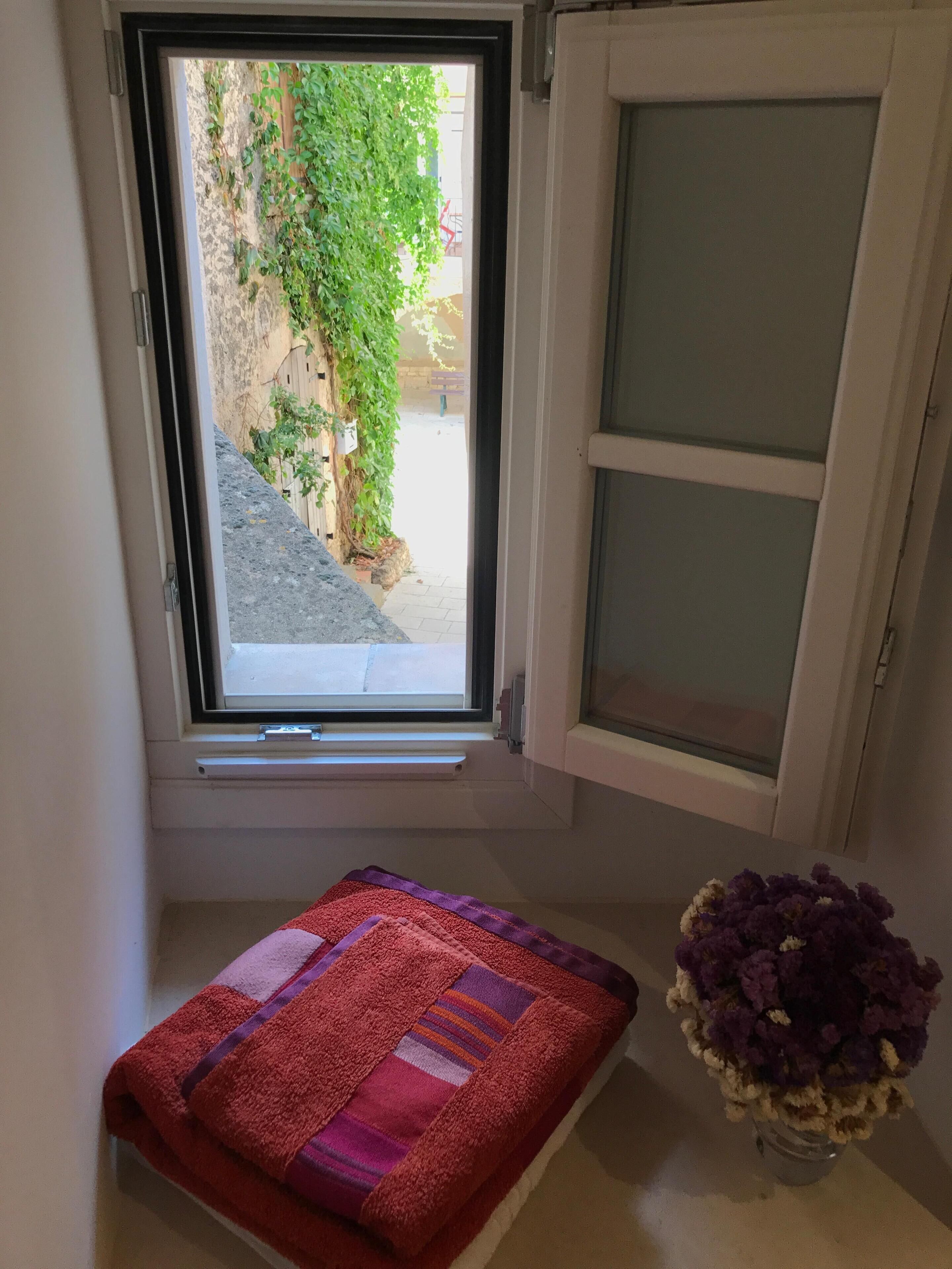 Ein Fenster mit Blick auf einen Garten, ein rotes und violettes Handtuch sowie eine Vase mit violetten Blumen.