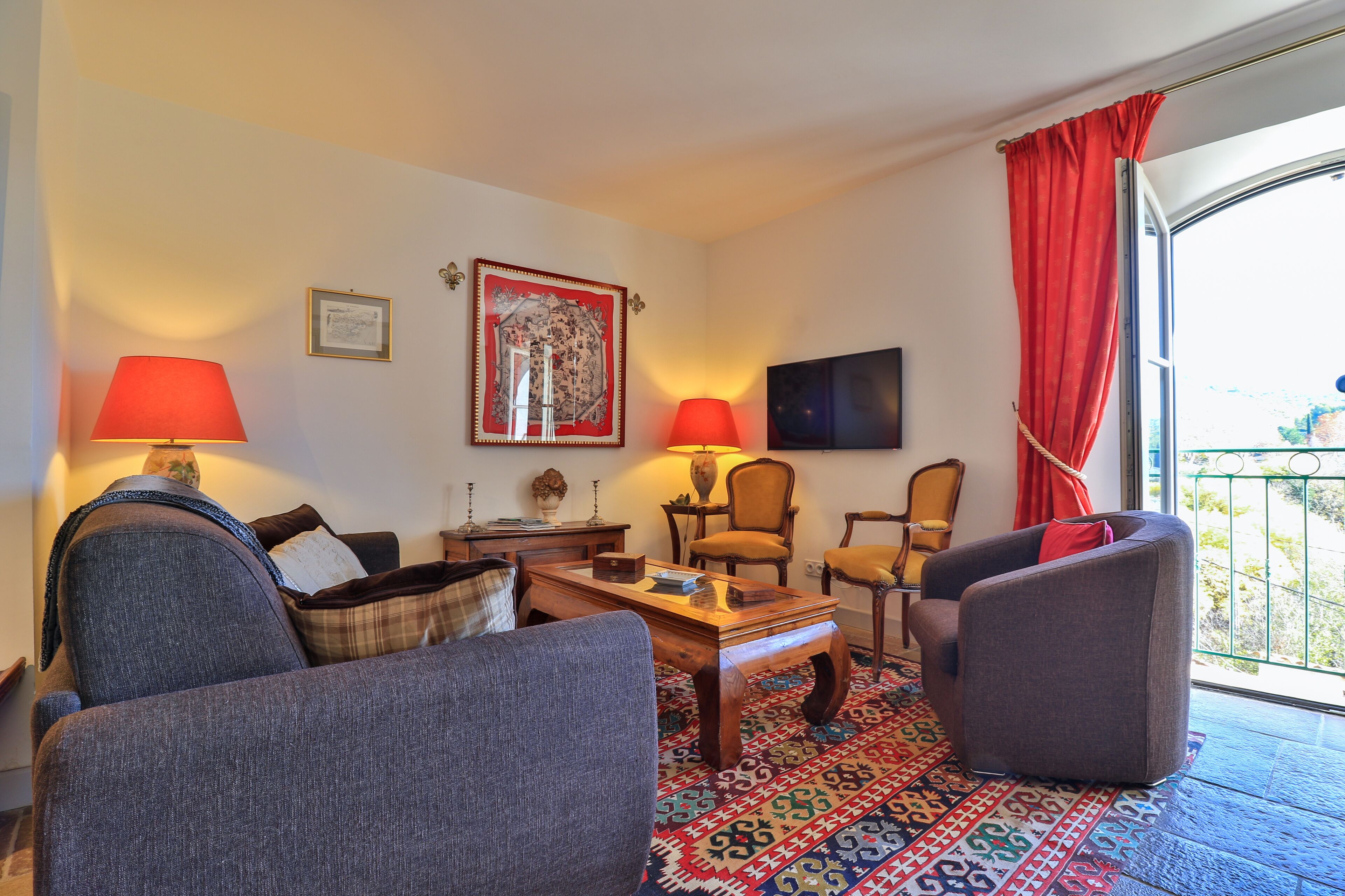 Uma sala de estar com sofá, poltronas, mesa de centro e duas luminárias. Um tapete estampado cobre o chão e há um quadro na parede.