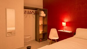 Ein Hotelzimmer mit einem Bett, einem Sessel, einem Nachttisch mit Lampe, einem Holzregal mit Handtüchern und einem Spiegel an der Wand.