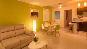 Ein Wohnzimmer mit einer Couch, einem Esstisch mit Stühlen, einem kleinen weißen Tisch mit Pflanze und einer Küchenzeile mit Schränken und Arbeitsfläche.