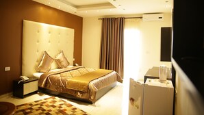 A hotel room with a large bed, a bedside table with a phone, a desk with a computer, and a refrigerator. - Kalaa Resort (Arnoun)