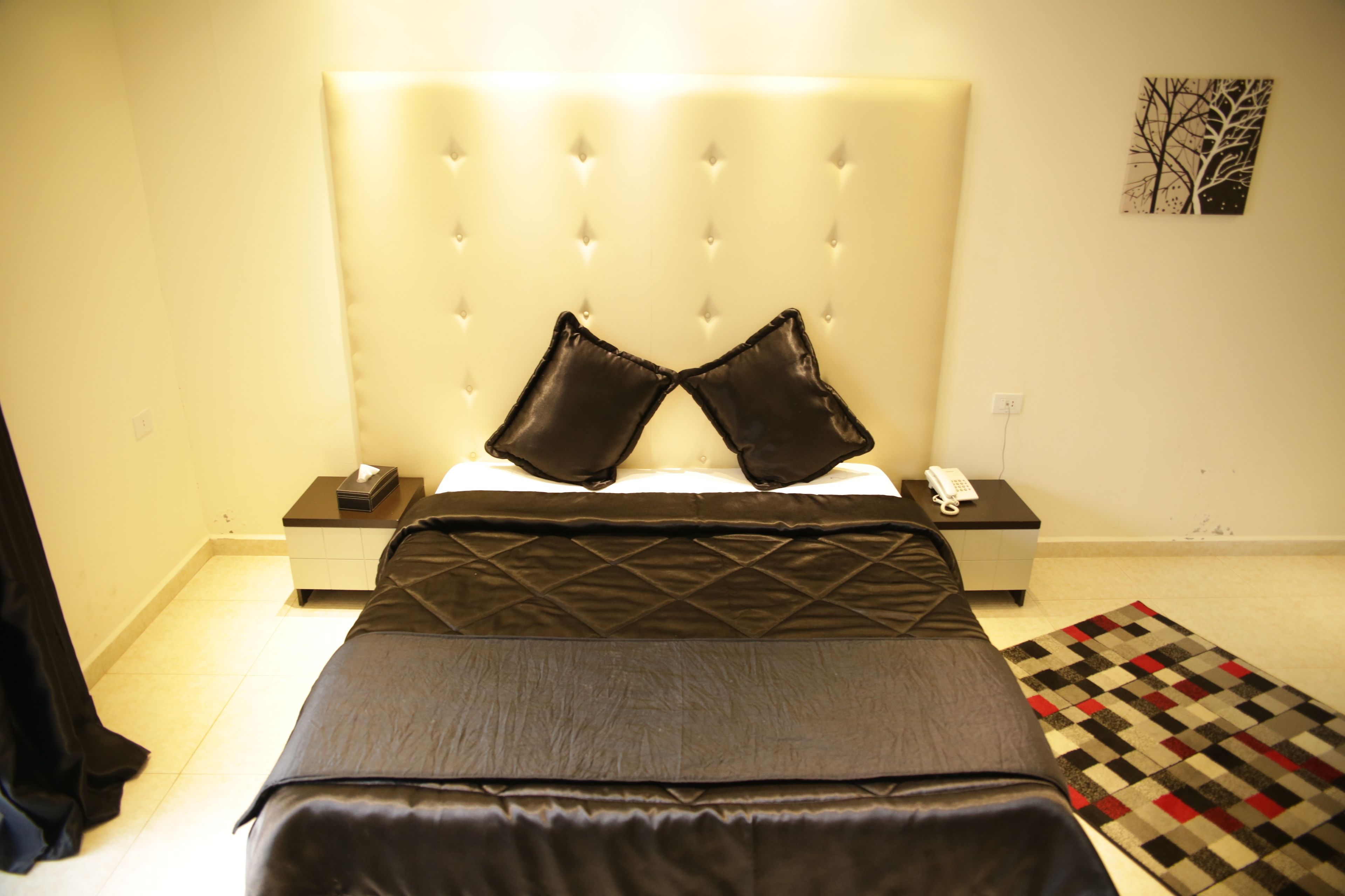 Ein Schlafzimmer mit einem gesteppten Kopfteil, einem Bett mit grauer Bettdecke, zwei schwarzen Kissen, einem Nachttisch mit Telefon und Lampe sowie einem gemusterten Teppich auf dem Boden.